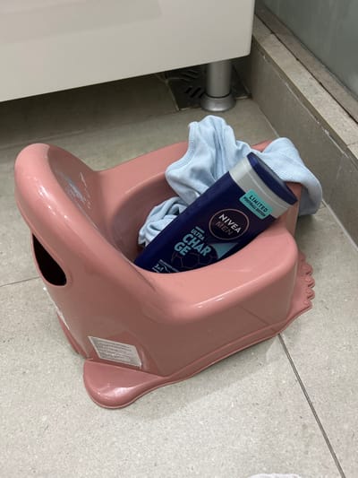 The scene, located indoors in Odorheiu Secuiesc, Romania, displays a pink plastic child's potty trainer positioned on a light-colored tiled floor. Within the potty, a dark blue Nivea Men "Ultra Charge 3in1" shower gel bottle, marked as a "Limited Football Edition," rests alongside a crumpled light blue fabric garment. The floor consists of large square tiles with visible grout lines. In the background, the lower portion of a white cabinet or fixture is visible, supported by two silver-colored cylindrical legs. A metal floor drain grate is present directly beneath the cabinet.