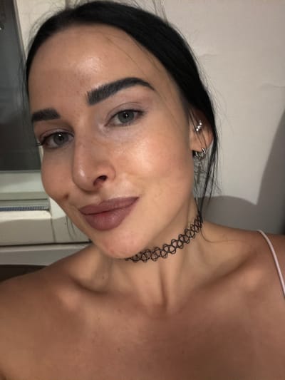 A close-up portrait shows a young woman with dark hair pulled back from her face, looking slightly upwards and to her left. Her head is tilted, giving the image a rotated perspective. She has prominent dark eyebrows and light-colored eyes, appearing to be a greenish-grey hue. Her lips are full, adorned with a matte, reddish-brown lipstick. Her skin appears fair with a few subtle marks or freckles visible.
She is wearing multiple silver-colored earrings on her visible left ear, including a helix piercing and a dangling earring with a small cross charm. Around her neck, she wears a black, stretchy, open-weave choker necklace, often referred to as a "tattoo choker." A thin white strap, possibly from an undergarment or top, is visible on her right shoulder. Her upper chest and shoulders are largely bare, indicating she may be wearing a strapless or thin-strapped top.
The background is a plain, light-colored surface, possibly a wall, with subtle variations in tone. One of the cropped images suggests a tiled surface, possibly hinting at a bathroom setting. The lighting is soft and even, suggesting an indoor environment with artificial light. The woman's expression is neutral, bordering on contemplative. The scene is set in Zanevka, Russia. No visible text is present in the image.