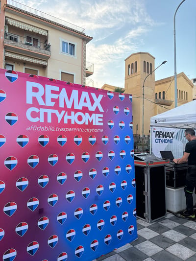 The image shows a marketing display for "REMAX CITYHOME" in Chianchitta, Italy. A large banner with a gradient color scheme (pink to blue) and repeated teardrop-shaped design stands in front of a building and a street. A man is partially visible, standing under a white tent branded with the "REMAX CITYHOME" logo, operating what seems to be audio equipment. The background features buildings with architectural details and a partly cloudy sky. The ground where the banner and tent are set up is paved with a checkerboard pattern. The additional text "affidabile.trasparente.cityhome" is also visible on the banner.