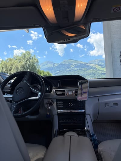 An interior view of a Mercedes-Benz car shows the dashboard, steering wheel, and passenger seat. The car is parked outdoors, with a scenic mountainous landscape visible through the windshield. The sky is blue with scattered clouds. A smartphone is mounted on the dashboard, displaying a navigation app with a route through Buchs (SG), Switzerland. The overall lighting suggests it is daytime. There are no people visible in the image.