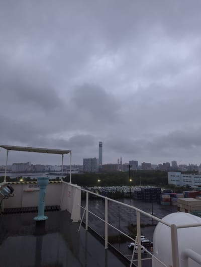 This is a photograph taken from a ship's deck, overlooking a port area in Chiba, Japan. The sky is overcast with dark clouds. In the foreground, part of the ship's deck is visible, including a white railing, a light blue bin, and a lamp. The deck surface is wet, indicating recent rain. Beyond the ship, a port facility is spread out, with numerous shipping containers stacked high, and rows of parked cars. A body of water separates the port from the city skyline in the background. The city features various buildings, including a tall, slender skyscraper and several other mid-rise structures. Trees line the area between the port and the city. The overall atmosphere is somber due to the cloudy weather.