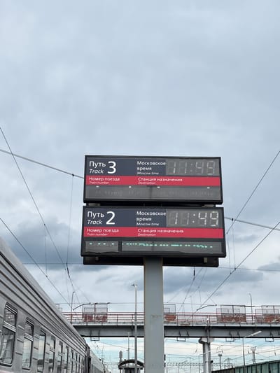 The image shows two digital display boards at a train station, likely in Povorino, Russia, under a cloudy sky. The boards display train information, including track numbers, train numbers, Moscow time, and destination stations. The top board shows "Track 3" and the bottom board shows "Track 2". Both boards display the Moscow time as "11:49". The destination display for both tracks is currently blank, indicated by a line of green lights. A portion of a grey train is visible on the left side of the image, running parallel to the display boards. In the background, a pedestrian bridge with railings and overhead electrical wires are visible, suggesting an active railway environment. The overall scene is one of a functional train station.