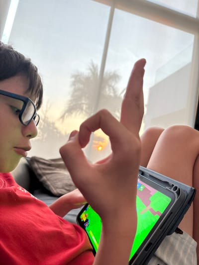 A person wearing glasses and a red shirt is looking upward toward a window. Their hand is raised, forming a circle with their index finger and thumb, framing the sunset visible through the window. The window also shows the silhouette of a palm tree. A phone or tablet in a black case is also visible. The screen of the device displays a game with green and brown graphics. Part of another person's leg is visible in the background.