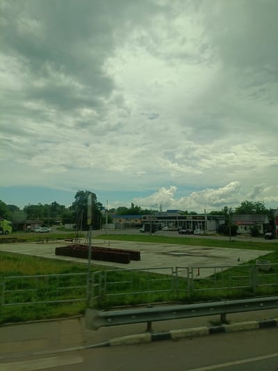 The image depicts an overcast sky with substantial cloud cover. In the foreground, a paved area with metal railings and a road barrier is visible, suggesting a roadside view. Beyond this, a grassy embankment leads to a large, open paved lot. Several structures are present, including a bright green truck, a small building with a red roof, and a row of commercial buildings. One of these buildings has signage that reads "WILDBERRIES". There are parked cars on the paved lot in front of the commercial buildings. In the middle ground, there are metal frames and what appear to be large, dark brown rectangular structures, possibly construction materials or storage units. The overall scene suggests an urban or suburban commercial and industrial area under a cloudy sky, likely in Saratovskaya, Russia, based on the provided context.