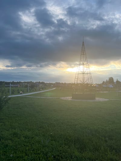 The image depicts a panoramic view of a park-like area in the city of Kamskiye Polyani, Russia, during what appears to be either sunrise or sunset, given the dramatic lighting. A tall, skeletal tower, likely an observation tower or a decorative structure, is prominently positioned in the mid-ground, silhouetted against the bright sky. The sky is filled with dark, heavy clouds that are illuminated by the sun breaking through in the distance, creating a dramatic contrast.
In the foreground, a lush green lawn covers the ground, with some scattered wildflowers. A paved pathway meanders through the scene, lined with streetlights and occasional benches, suggesting a well-maintained public space designed for recreation. Beyond the pathway, a scattering of residential buildings and trees can be observed, indicating the proximity of a residential area to the park. The overall atmosphere is one of natural beauty enhanced by human design, with the dramatic sky adding a touch of moodiness to the scene.