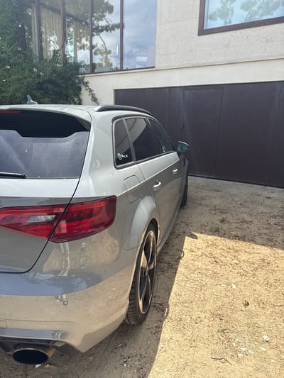 A light grey hatchback car is parked on a sandy or gravelly surface next to a modern building. The image is oriented with the car's right side and rear facing the viewer. The ground is dry, strewn with small stones, sparse weeds, and some debris like pine needles. Strong sunlight creates distinct shadows, indicating a clear, sunny day.
The car features a sleek design with dark-tinted rear windows and a prominent, large black exhaust pipe visible on the right side of the rear bumper. It has black, multi-spoke wheels with low-profile tires. The rear taillights are red, and a circular parking sensor is visible on the light grey bumper. The car appears somewhat dusty. A black roof spoiler is present above the rear window. On the rear side window, a sticker displays the text "@Min_cli" and a small skull-like graphic underneath it.
To the left, a modern building with a light-colored, possibly stone or concrete, facade is visible. It features large, dark-framed windows that reflect tall trees and the sky. Green foliage or climbing plants grow along the base of the building, below the lower windows. A dark-colored wall or garage door is seen further back. The overall scene suggests a private residence in a sunny, possibly coastal or semi-rural environment, consistent with Lège-Cap-Ferret, France. No people or activity are present.