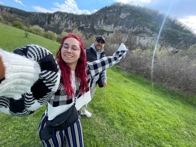 A selfie-style photograph shows two people in an outdoor setting, presumably taken in the vicinity of Barkach, Bulgaria. The image is oriented sideways. A woman with red hair, wearing glasses, is closer to the camera and smiling. She is wearing a black-and-white checkered sweater. To her left, a man in a gray jacket and black hat is also smiling. In the background, there is a large rock face, green fields, and a blue sky with white clouds. The woman is also wearing striped pants and a black bag. There is a flash of light on the rock face. The photograph is taken at an angle, showing the environment as a backdrop.