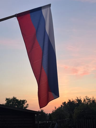 The image depicts the Russian flag hanging from a flagpole against a sky at dusk. The flag is predominantly red, blue, and white, arranged in vertical stripes. The sky exhibits a gradient of colors, transitioning from a pale blue at the top to soft oranges and pinks near the horizon, suggesting either sunrise or sunset. In the background, silhouetted trees and a dark, low-lying structure, possibly a shed or fence, are visible. The scene appears to be an outdoor setting, likely a residential or rural area, with no people present. The overall atmosphere is serene and quiet.