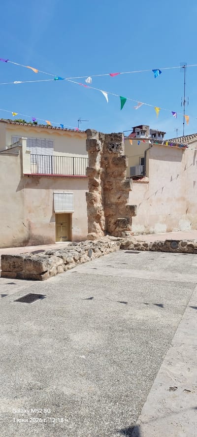 The image depicts a narrow street in Canals, Spain. Strings of triangular pennants are strung overhead against a clear blue sky. The facade of a building lines the left side of the street, featuring a mix of architectural styles, including aged stucco, stone remnants, and a balcony. A pathway is visible to the right.