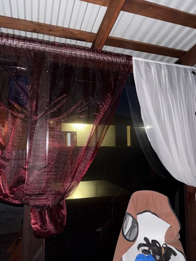An exterior shot taken at night in the city of Stolbishche, Russia shows a view through a covered patio or gazebo. Two curtains, one dark red and sheer, the other white and opaque, hang from a wooden beam that supports a corrugated metal roof. Through the gap between the curtains, a brick building with illuminated windows is visible in the distance. A partially visible object, possibly a chair or stool covered with a brown fabric and a blue plastic bag on top, is in the foreground on the right. The scene is dark, with the interior of the building and the foreground objects being the main sources of light.