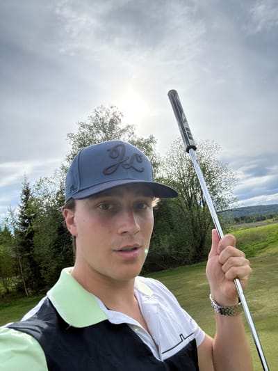 A man holds a golf club overhead in a vertical, low-angle selfie taken outdoors, likely on a golf course. He wears a blue baseball cap with a dark emblem, a white shirt with a light green collar, and a metal watch on his left wrist. Trees and a grassy field are visible in the background, along with a bright sky. The location may be Hakadal, Norway.