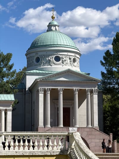 The image shows a view of the Church of the Archangel Michael in Arkhangel’skoye Estate, Russia. The church is a neoclassical structure with a prominent green dome topped by a cross. The facade features classical columns and decorative friezes. In the foreground, a white balustrade overlooks a stone staircase leading to the church entrance. Two women are visible on the staircase, one holding a smartphone, possibly taking a photograph. Tall evergreen trees frame the right side of the building. The sky is blue with scattered white clouds.