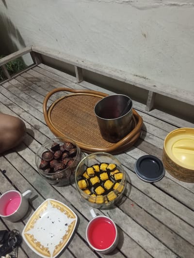Scene: An outdoor setting in Pulokiton, Indonesia.
Objects:
* A light-colored wooden deck.
* Two bowls of cookies: one with chocolate cookies and sprinkles, the other with yellow-colored cookies.
* A woven tray holding a stainless steel container.
* Two mugs: one with pink liquid, one with red liquid.
* A ceramic plate with gold trim.
* A yellow container with a dark gray lid.
* A set of keys.
People:
* A person's bare leg is visible in the left side of the frame.
Actions:
* No actions directly observable.
Interactions:
* Items are arranged on the deck.
Environment:
* The setting is lit.
* A white wall is behind the deck.