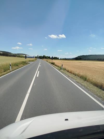 A perspective shot from a vehicle shows a two-lane asphalt road leading towards a distant intersection. The road is flanked by fields of dry, golden crops on either side, suggesting late summer or early autumn. The sky is a vibrant blue with scattered white clouds and a faint contrail. Road markers with black diamonds and white circles are visible along the roadside. In the distance, a modern building with a flat roof and a tall spire, possibly a church or industrial structure, is visible. A slight blur across the bottom of the image indicates the presence of a vehicle's interior, likely a dashboard or windshield. The location is on the outskirts of Lauda-Königshofen, Germany, specifically on a rural road that leads into the town, given the agricultural landscape and the distant architecture typical of the region.