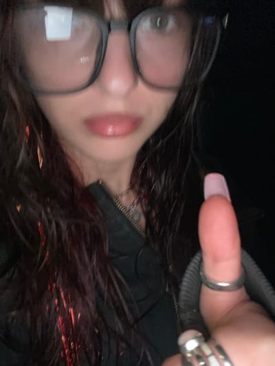A blurry, close-up selfie shows a person with dark, wavy hair wearing glasses. Their lips are glossy. They have a light pink manicure and wear rings on their fingers. They appear to be wearing a dark jacket with a zipper. The picture is taken from a slightly high angle, creating a distorted perspective. Reflections obscure the glasses lenses. The background is dark and undefined.