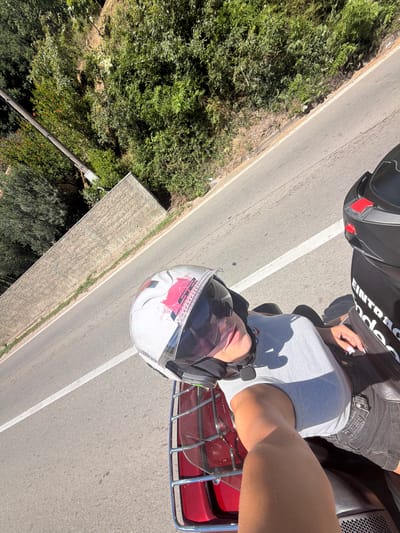 An overhead shot captures a person riding a scooter along a paved road in Mišići, Montenegro. The rider, wearing a white helmet with pink and black accents and sunglasses, is seen from the chest up. They are wearing a light gray t-shirt and black shorts. Sunlight casts a shadow on the rider and the road. To the right of the rider, another scooter helmet is visible. The environment consists of a road bordered by lush green vegetation and a low, textured stone wall on the left. The sky is a clear blue.