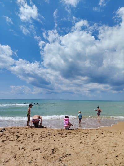 The image captures a sunny day at a beach in Blagoveshchenskaya, Russia. The sky is a vibrant blue, dotted with fluffy white clouds, suggesting fair weather. The sea is a tranquil turquoise, with gentle waves lapping at the sandy shore.
In the foreground, three children are visible. One child, wearing a pink life vest, is seated on the sand, facing the water. Another child, wearing a blue shirt and shorts, stands in the shallow water, looking towards the sea. A third child, in colorful swim trunks, is bent over, actively digging in the sand. A young boy, with dark hair and wearing swim trunks, stands on the beach near the water's edge, observing the scene.
In the background, several people are swimming and playing in the ocean. A woman in a patterned bikini is also standing in the water, facing away from the camera with her arm outstretched. The overall atmosphere is one of leisure and enjoyment, with people taking advantage of the beautiful weather to relax and have fun at the beach. The time of day appears to be late morning or early afternoon, given the bright sunlight. There is no visible text in the image.