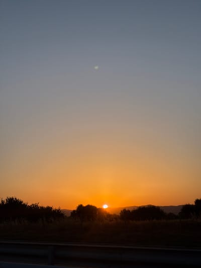 The image depicts a sunset over a landscape. The sky transitions from a pale blue at the top to a warm orange near the horizon. A faint white orb, possibly the moon or a lenticular cloud, is visible in the upper sky. The horizon is lined with dark silhouettes of trees and vegetation. In the distance, rolling hills are dimly visible against the bright sky. A dark, horizontal structure, likely a road barrier or railing, runs across the foreground. The overall scene is serene and captures the natural beauty of the twilight. Without additional information, the specific location within Kolarovo, Bulgaria cannot be definitively identified.