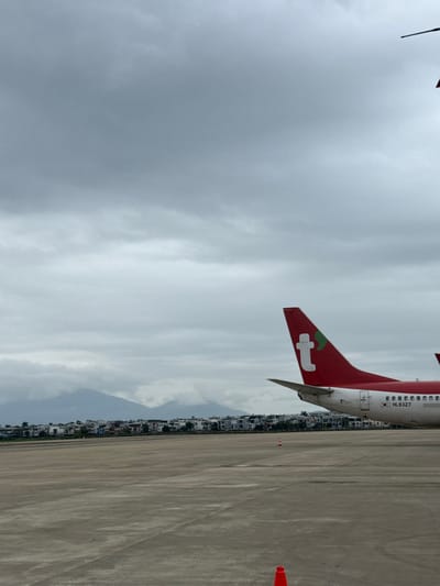 The scene depicts an airport apron under an overcast, grey sky, located in Hòa Cường Ward, Vietnam. A red and white aircraft is partially visible on the right side, showing its tail fin and a section of its fuselage. The red tail features a white 't' logo with a small green leaf. The white fuselage displays a South Korean flag decal and the registration number "HL8327". The foreground is a vast expanse of grey concrete pavement. In the mid-ground, beyond the tarmac, a dense residential area with numerous low-rise buildings extends horizontally, backed by mountains partially obscured by clouds. Two orange traffic cones are present on the tarmac; one in the lower right foreground and another positioned further back. No individuals are visible.