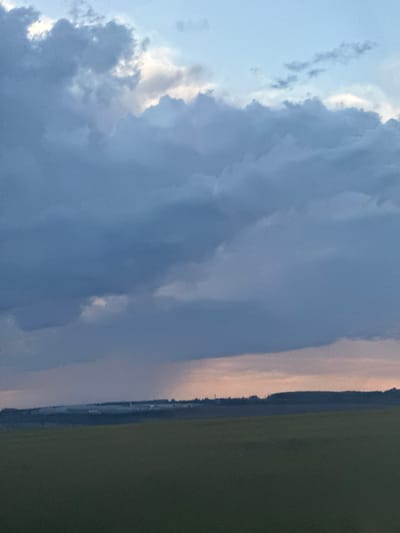 The image displays a panoramic view of a rural landscape under a dramatic sky. The foreground is dominated by a vast, dark green field. Beyond this field, a gentle slope rises, lined with a dark silhouette of trees and low-lying structures, suggestive of a small settlement or industrial area. The sky is filled with large, heavy, dark blue clouds, with patches of lighter grey and white indicating breaks or thinner cloud formations. A faint, misty rain shower is visible in the mid-ground, creating a subtle veil over the landscape. The horizon line is low, emphasizing the expansive sky. The location is identified as Пермяки, Russia, a rural area. There are no people or specific actions depicted in the image; it is a static environmental scene.