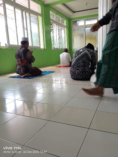 Three men pray in a room with white-tiled floors, green walls, and bright windows. The first man wears a batik shirt and a white cap. The second has on a patterned top and black head covering. The third wears plaid and green striped pants. They are all kneeling on the floor. In the foreground, a barefoot man is walking past. A watermark is visible at the bottom of the image.