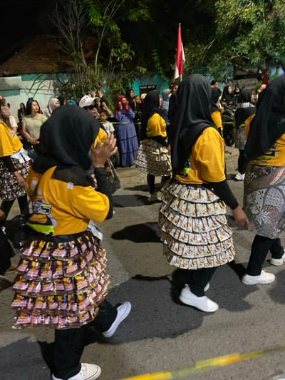 This image depicts an outdoor night scene in Kedungbanteng, Indonesia, likely a community parade or event. In the mid-ground and foreground, several individuals, predominantly women, are walking in formation, largely with their backs to the camera. They wear yellow short-sleeved shirts over dark long-sleeved undershirts, black trousers, and white sneakers. A distinctive element of their attire consists of multi-tiered skirts constructed from numerous small, individual product packages; two discernible types include reddish-pink and gold snack wrappers and lighter brown and beige instant coffee sachets. Many participants wear dark headscarves. One person in the immediate foreground has their right arm raised. In the background, a crowd of spectators lines the left side of the street, observing the procession. An Indonesian flag, red and white, is visible on a pole further back, against a backdrop of dark foliage and dimly lit building structures. The paved ground shows distinct shadows, indicating illumination by artificial light sources during nighttime.