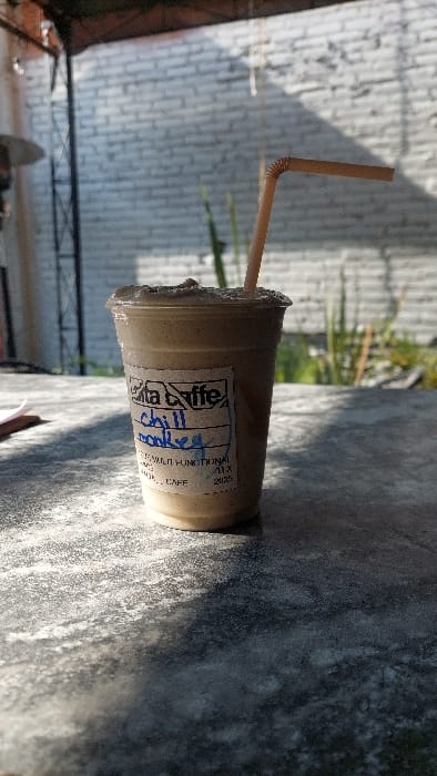 The image depicts a close-up, static view of a plastic beverage cup in an outdoor or semi-outdoor commercial setting in Tlaxcala City, Mexico. The central object is a clear plastic cup containing a light brown, frothy, blended beverage, with a light brown, bent plastic straw inserted. A white rectangular label affixed to the cup displays "Vita Caffe" at the top and the handwritten blue text "chill monkey". Below this, partially visible text includes "SABANA HUNGARINA TLX" and "CAFE", specifically identifying the location as a Vita Caffe branch on Sabana Húngara street in Tlaxcala City. The cup rests on a dark grey, textured surface, likely a tabletop, with strong, directional sunlight creating sharp shadow patterns. In the background, a light-colored brick wall is visible, with minimal green foliage behind the cup. A dark metal structural element is partially visible in the upper left, suggesting an awning or canopy overhead. No individuals, actions, or direct interactions are present in the scene.