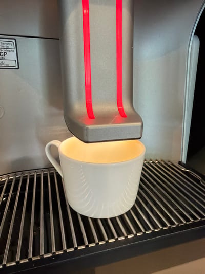 This is a close-up, overhead shot of a white ceramic mug with a ridged texture, positioned to receive liquid from a silver machine. The mug is tilted, with its opening facing a dispensing spout. Inside the mug, a warm, yellow light emanates, suggesting the liquid being dispensed is hot. The machine has a sleek, modern design with two horizontal red LED lights on the dispensing unit, which appears to be made of brushed metal. To the right of the mug and machine, a dark metal grate is visible, likely part of the appliance's design or function. A small label with text is partially visible in the lower left corner of the image, but the text is too small and out of focus to be legible. The overall scene suggests a coffee or beverage machine in operation, likely in a modern setting such as a kitchen or café. The lighting is warm and inviting, and the focus is on the interaction between the machine and the mug. There are no people visible in the image. The image appears to have been taken indoors.