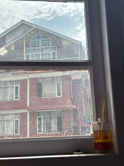 The image shows a view through a dirty window frame, looking out at two brick buildings. The top window frame shows the gable end of a building with a large, multi-paned window with a teal-colored tint. Below this, the lower window frame shows the facade of another brick building with multiple windows, each covered with white curtains. The scene appears to be in an urban environment. On the windowsill, to the right, is a clear glass diffuser bottle filled with amber-colored liquid and several wooden reeds. A small black incense holder is also visible on the windowsill. The windows are noticeably smudged and streaked. The location is identified as Chichoga, India.