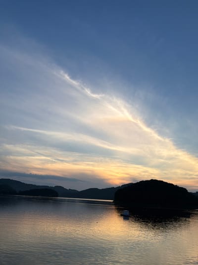 This image depicts a tranquil sunset over the Rożnowskie Lake in Gródek nad Dunajcem, Poland. The sky transitions from a deep blue at the top to hues of orange and yellow near the horizon, streaked with wispy white clouds. Silhouetted hills form the backdrop, with a prominent, darkly shaded island in the foreground. The lake's surface is calm, reflecting the sky's colors and creating subtle ripples. A small, white, box-like structure floats on the water near the island. The overall scene conveys a sense of peace and natural beauty.