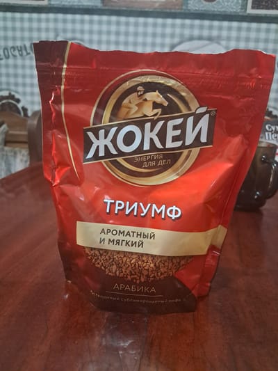 A close-up, eye-level shot captures a red coffee bag with gold accents placed on a polished wooden table. The bag features prominent Cyrillic lettering, the brand name "ЖОКЕЙ" (JOCKEY) stylized over an image of a jockey on a horse, and a subtitle "ТРИУМФ" (TRIUMPH). Below the title, smaller text reads "АРОМАТНЫЙ И МЯГКИЙ" (AROMATIC AND SOFT). The bottom portion of the bag shows a texture of coffee granules, with the word "АРАБИКА" (ARABICA) above the fine print "Растворимый сублимированный кофе" (Instant sublimated coffee). To the right, a dark-colored mug with a handle is partially visible, suggesting it contains a beverage. The background is a checkered fabric with light and dark green squares, and a portion of what appears to be a shelf with other items is visible in the upper left corner. The lighting is moderate, casting some soft shadows. The image was taken in Chobruchi, Moldova.