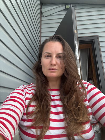 This is a vertical, medium close-up selfie of a woman with long, brown hair and brown eyes. She is wearing a red and white striped long-sleeved shirt. Her gaze is directed towards the camera, with a neutral expression.
The background consists of a grey, horizontal wooden wall. To the left of the woman is a dark grey door frame, with a portion of the door open, revealing a glimpse of what appears to be an interior space. Above the door frame, there are some metal fixtures, including what looks like a label with text that is too blurry to read. To the far left, a black outdoor light fixture is visible.
The scene appears to be outdoors, possibly on a porch or deck given the siding and the presence of the door. The lighting suggests it could be daytime, but there are no strong shadows that would indicate a specific time. The weather is unclear.
The woman's hair is slightly messy and falls over her shoulders and chest. Her skin tone is fair. The overall impression is a casual, everyday moment captured in a selfie.
The provided location context is "Буран Снт, Russia." There is no discernible text in the image that is legible.