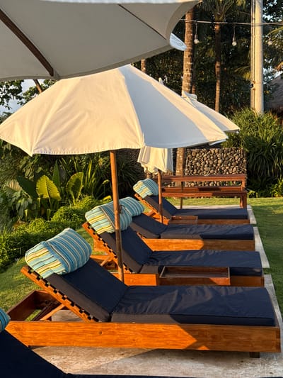 The image depicts a luxury outdoor resort setting in Medahan, Indonesia. Several lounge chairs with dark blue cushions and striped blue/green pillows are arranged in a row. Each lounge chair is made of wooden planks, and they are positioned on a concrete surface next to a lawn. White sun umbrellas are present, providing shade. Tropical foliage and trees surround the area. A wall of black pebbles or rocks can be seen behind the chairs. Overhead lighting is visible above the trees. The scene is bathed in sunlight.