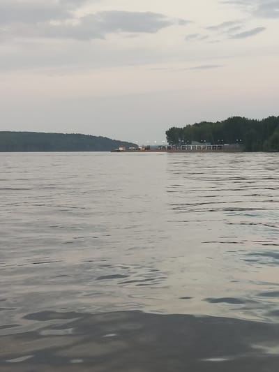 The image depicts a wide, calm river with gentle ripples. In the distance, a wooded shoreline is visible, with a long, low-lying structure situated along the water's edge, featuring white posts and a roofed area. A barge or a similar vessel with cargo is moored near this structure. The sky is overcast with soft, diffused light, suggesting either early morning or late evening. The overall scene is serene and tranquil, conveying a sense of natural beauty and quietude. The location is identified as Galovo, Russia, implying this is a view of the river within or near that city.