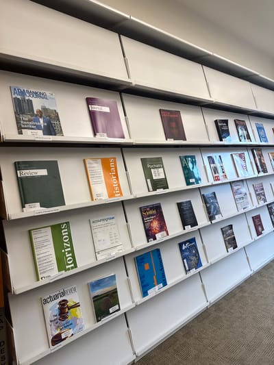The scene depicts an interior view of the Bentley Library at Bentley University in Waltham, Massachusetts, United States. White, multi-tiered display shelves are affixed to a white wall, presenting numerous academic journals and magazines face-out. The shelves are arranged in horizontal rows and columns, creating a grid-like pattern. Visible titles include "ABA Banking Journal," "Academy of Management Journal," "American Cinematographer," "Academy of Management Review," "historians," "The American Journal of Psychiatry," "Horizons," "accounting today," "American Scientist," "actuarial REVIEW," "Icons of American Business," and "American Sociological Review." Many displayed items feature small white labels, some clearly marked "BENTLEY LIBRARY." The floor is covered with patterned beige and brown carpet. No individuals are present in the frame. The area is uniformly lit, with light reflecting off the white surfaces.