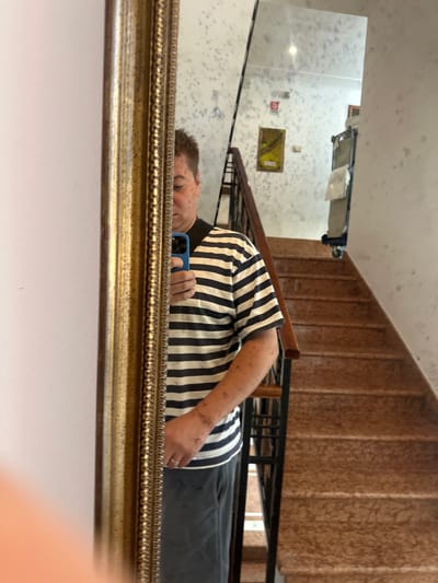 A person is taking a mirror selfie in a building, likely an apartment complex or hotel, in San Martino Buon Albergo, Italy. The person is wearing a black and white striped shirt and gray pants. They are holding a blue smartphone in their right hand. The mirror is ornate and gold-colored. In the reflection, a staircase with marble steps and a black railing is visible. The walls have a textured pattern with blue and white splotches. There is a framed picture on the wall above the staircase. The overall environment suggests an interior common area or hallway.