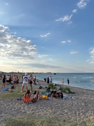 The image depicts a crowded beach scene on a sunny day in Kastrup, Denmark. A multitude of people are scattered across the sandy shore, engaged in various activities such as socializing, lounging, and walking. Several groups are picnicking on blankets, with food and drinks laid out. In the background, the calm blue water of the sea stretches towards the horizon, where a line of wind turbines is visible. The sky is a clear blue with scattered white clouds. The overall atmosphere appears relaxed and festive, indicative of a popular summer gathering spot.