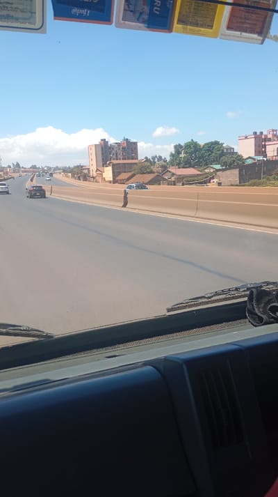 A view from inside a vehicle, likely a car, driving on a highway. The road is wide with a central concrete barrier. Buildings and trees are visible beyond the barrier. Cars are seen on the highway. Windshield wipers and a portion of the dashboard are visible. The sky is blue with some clouds. Cards or papers are affixed to the top of the windshield.