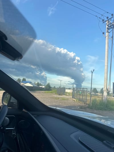 A wide shot captures a plume of smoke rising into a bright blue sky, viewed through the windshield of a vehicle. The smoke plume is large and billows upwards, appearing white and grey against the clear sky. In the foreground, the dashboard of the vehicle is visible, along with a side mirror. Beyond the vehicle, the landscape consists of a dirt road, some houses with blue roofs, a wooden fence, and tall grass. A power pole with wires stands prominently on the right side of the image. The overall scene suggests an industrial or natural event causing the smoke. The location is Oranzhereika, Russia.