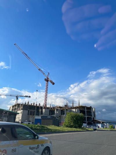 The image shows a construction site in Bittsa, Russia. A multi-story building is under construction, with several cranes visible. A taxi cab is parked on the side of the road next to the construction site. The sky is blue with scattered clouds.