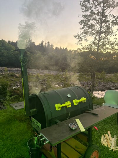 A green barbecue smoker is positioned on a grassy area near a rocky river. Smoke billows from the smoker's chimney and the main chamber. A Texas license plate with the number "CPG-2985" is affixed to the smoker. A metal bucket hangs from the smoker's side. The scene is illuminated by the soft light of either dawn or dusk, with trees and a hazy sky in the background. The location is described as Kapreshumi, Georgia.