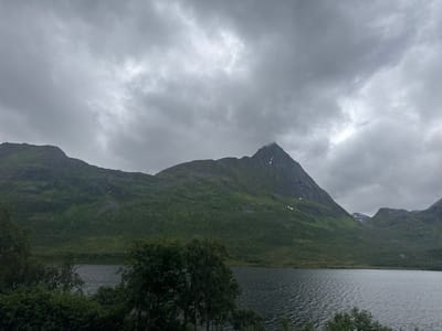 The image depicts a dramatic, overcast landscape in Gullesfjord, Norway. A dark, choppy lake dominates the foreground, with the choppy surface suggesting a brisk breeze. The lake is framed by lush green trees and foliage in the lower part of the image.
Behind the trees and lake, a series of imposing, verdant mountains rise steeply. The slopes are covered in dense vegetation, appearing as a carpet of green. Patches of snow or light-colored rock are visible on the higher, steeper inclines of the central, most prominent peak. This peak is sharp and imposing, with a small cloud or mist clinging to its summit, adding to the atmospheric and somewhat mysterious feel of the scene. The sky is heavily clouded, with large, grey, and white formations that suggest a cloudy or potentially rainy day. The light is diffused, creating a somber and moody ambiance.
The scene is a natural, mountainous landscape, devoid of any visible people or man-made objects, except for what appear to be distant power lines or pylons on the right side of the image, partially obscured by the terrain. The overall impression is one of raw, untamed natural beauty, with a sense of solitude and the power of nature. The time of day is not clearly discernible due to the overcast conditions, but the general mood leans towards a cool, possibly late afternoon or early morning in a temperate climate. There is no visible text in the image.