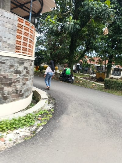 The image portrays a semi-urban or rural road scene in Cangkuang Kulon, Indonesia. The paved asphalt road, which curves gently to the right, is flanked by a concrete drainage culvert on its left edge and dense green foliage on the right. In the immediate left foreground, a distinctive rounded structure features a base of stacked stone veneer and an upper section of terracotta-colored breeze blocks, surmounted by a partial metallic canopy. A female individual, clad in a light-colored long-sleeved shirt, blue jeans, and a black hijab, is walking away from the camera on the left side of the road, wearing dark flip-flops. Further down the road on the right, a second individual, wearing a bright green vest over a long-sleeved garment, is seated on a black scooter, also facing away from the camera. The background includes numerous mature green trees, fallen leaves scattered on the ground, and several residential buildings with visible orange-tiled roofs. A yellow bin or cart is situated on the right side of the road among the foliage.