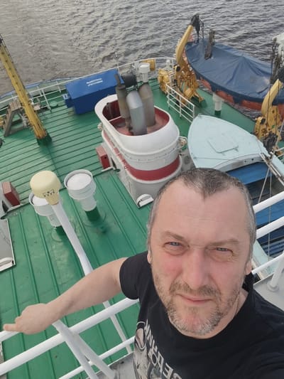 A middle-aged man with graying hair and a beard is taking a selfie on the deck of a ship. The ship's deck is painted green and features various nautical equipment, including a red and white structure with metal chimneys, white vents, a blue box, and a covered lifeboat secured by yellow cranes. The man is wearing a black t-shirt and is holding onto a white railing with his left hand. Beyond the ship, there is a view of water. The location is possibly near Novodevich'e, Russia, given the context of the prompt.