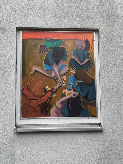 The image shows a painting displayed in a window on a textured grey building wall. The painting depicts five people sitting on the floor, playing cards. The background of the painting is a warm gradient of orange to brown. In the upper left corner of the painting, there are a pair of brown shoes and a green bag. The people are rendered in a somewhat abstract, impressionistic style with bold brushstrokes. One person, wearing an orange top, is in the foreground, their back to the viewer, with their head tilted down towards the cards. Other figures are also engaged in the game, with some looking at their cards and others observing the game. A blue water bottle is visible on the floor. The painting is framed by a white border and installed in what appears to be a window. The building exterior is rough and uneven.