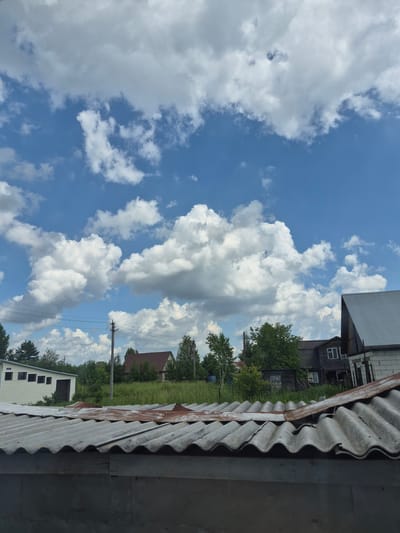 The image captures a view of a residential area under a bright, cloudy sky. In the foreground, the corrugated roof of a building, likely a garage or shed, is visible, showing signs of age and some rust. Behind it, a row of houses and other structures are set against a backdrop of lush green trees and grass. Power lines stretch across the scene, supported by a utility pole. The sky is a vibrant blue, filled with large, puffy white clouds, characteristic of a summer day. The architectural style of the houses suggests a rural or semi-rural setting in Sokur, Russia, with older wooden buildings and a more modern brick structure visible. There are no people or animals present in the image.