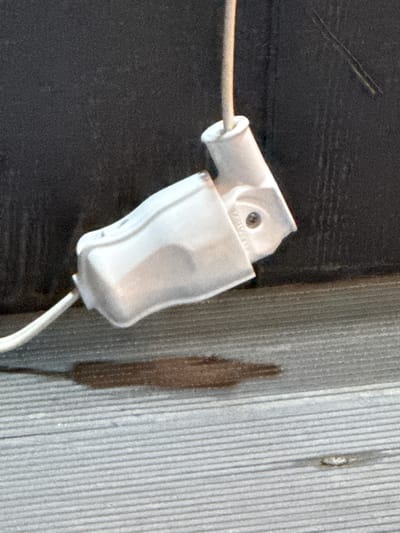 A white electrical plug is hanging down from an unseen source, attached to a white cord. The plug is angled, with the female receptacle facing upwards and slightly to the right. The cord emerges from the bottom left of the plug and extends out of frame. The background is a dark, textured surface, possibly wood or siding. The plug is positioned above a light gray, slatted surface, resembling a deck or boardwalk. A dark, damp stain is visible on the slatted surface directly beneath the plug. The overall scene appears to be outdoors.