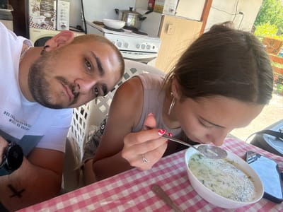 The image depicts two individuals, a male and a female, seated at a checkered table in what appears to be a semi-outdoor kitchen and dining area in Staro Selo, Bulgaria.
The male, positioned on the left, has short blonde hair, a beard, and a mustache. He wears a white textured polo shirt, a gold chain necklace, and a black razor blade-shaped earring in his left ear. A black smartwatch is visible on his left wrist, along with a dark cross tattoo on his left forearm. He is looking directly towards the camera.
The female, positioned on the right, has dark brown hair pulled back. She wears a light-colored sleeveless top and grey denim shorts are partially visible beneath the table. She has silver hoop earrings in her left ear, and a silver ring with a small charm on her left ring finger. Her fingernails are painted with a red and white design. She is actively eating, holding a silver spoon with food to her mouth from a white oval bowl containing a light-colored soup with green herbs, consistent with Tarator. A black smartphone is resting face-up on the tablecloth to the right of her bowl.
The table is covered with a red and white checkered tablecloth. In the background, a kitchen area is visible with a white two-burner electric stove, a pot, and a white bowl on it. A white PROLUX microwave oven is situated to the left of the stove. Above the microwave, two cardboard packages with Bulgarian text are visible, one featuring grapes and mentioning "Thracian wine" ("тракийска вино") and the other mentioning "Bulgarian garlic" ("български чесън"). To the right, a wooden cabinet or door frame is present, along with an electrical outlet and cable. Beyond the dining area, lush green foliage and bright sunlight indicate an outdoor setting during daytime, with a wooden railing partially visible. White plastic chairs with a lattice backrest are also partially visible.