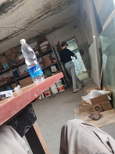 The image depicts an indoor scene, possibly a storage area or workshop, in Sokuluk, Kyrgyzstan. In the foreground, a person's legs and lap are visible, covered in light-colored fabric, suggesting they are seated. To the left, a portion of a pink wooden surface, possibly a table or workbench, is prominent. On this surface rests a clear plastic bottle of "Legend" brand water.

In the mid-ground, a person wearing a dark t-shirt, a cap, and light-colored jeans is walking away from the camera towards an open doorway. The doorway leads to a room with checkered blue and white walls. The background reveals shelves filled with various boxes and containers, indicative of storage. The environment appears somewhat cluttered and utilitarian, with a concrete floor and a textured, possibly damaged, ceiling. A pair of work gloves is visible on a wooden board in the lower right portion of the image. The overall impression is a mundane, everyday scene within a functional space.