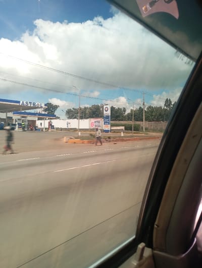 This image depicts a roadside scene in Sigona ward, Kenya, viewed from inside a vehicle through its right-side window, with the window frame and interior trim visible on the right. The sky is partially blue with large white cumulus clouds, traversed by multiple overhead power lines and several streetlights.
A multi-lane asphalt road with white lane markings occupies the foreground and mid-ground. A concrete median with orange-painted curbs and minimal vegetation separates the road lanes.
On the far side of the road, a blue and white "ASTROL" petrol station is visible, featuring a canopy over fuel pumps. A tall, blue and white "ASTROL" sign with a star logo stands prominently near the station entrance, alongside a smaller red and white sign displaying the number "6". Behind the station, a light-colored wall or fence is backed by a line of green trees and bushes.
Two motion-blurred individuals are visible walking on the far side of the road near the median; one appears to be moving left, and the other right. Additional indistinct figures are present near the petrol pumps at the "ASTROL" station.